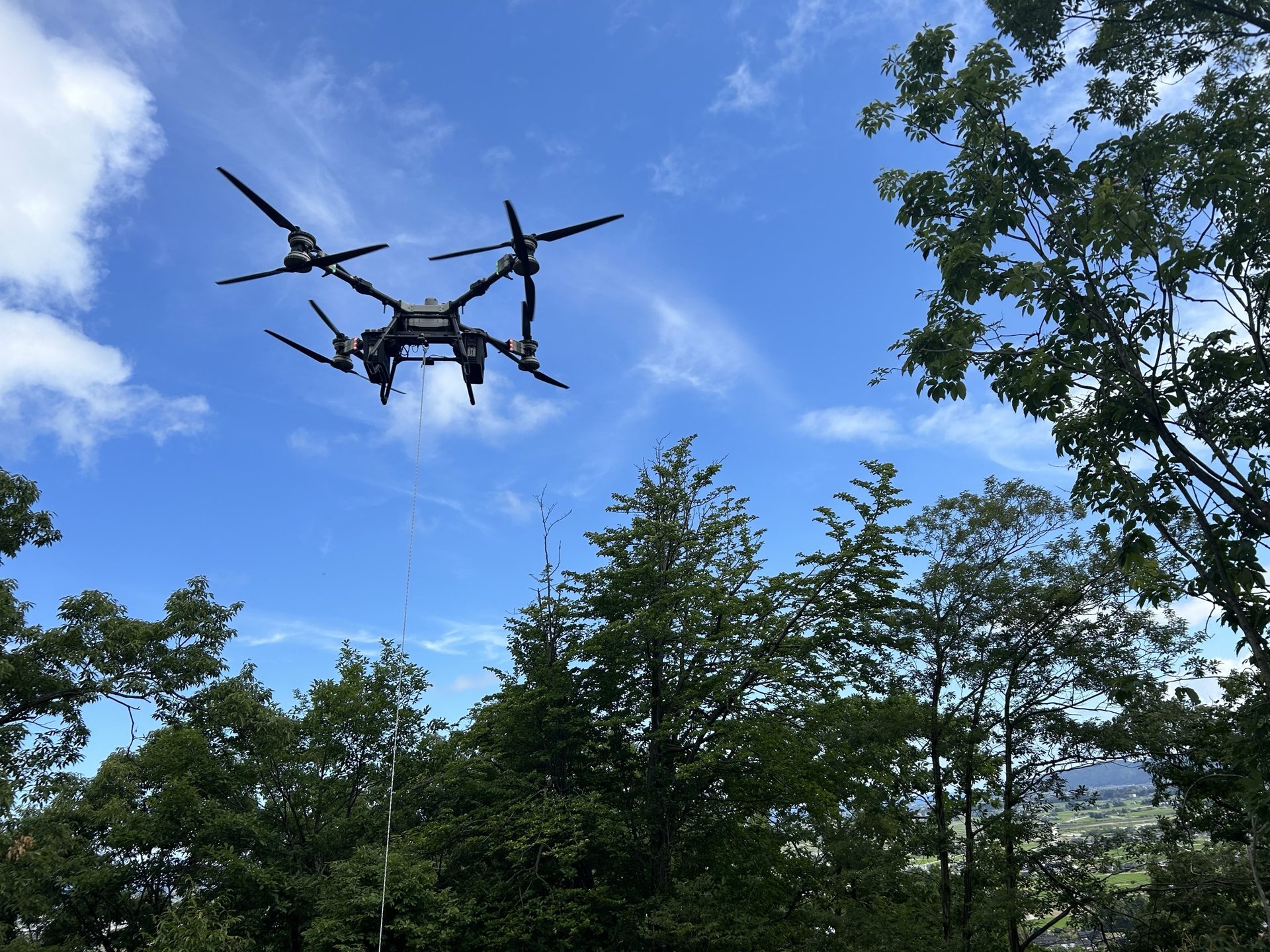 山間部の森林上空を飛行し資材を吊り下げ運搬するドローン